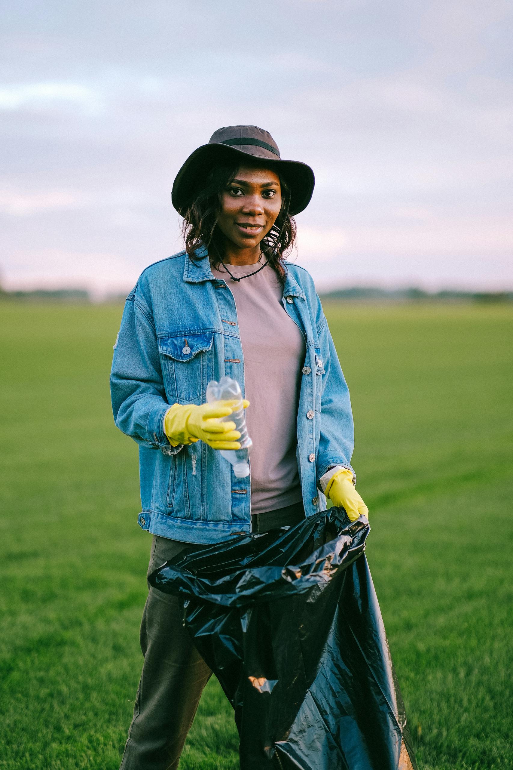 A woman in a denim jacket collecting trash in a green field, promoting volunteer work.